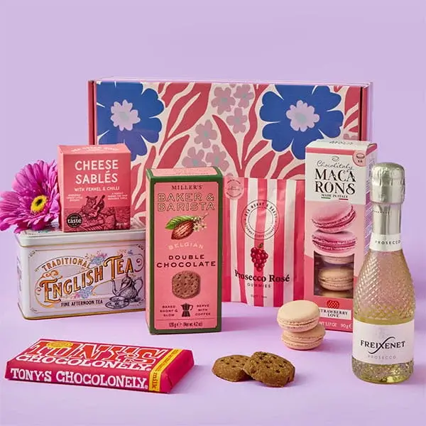 A colourful gift hamper featuring sweet treats, biscuits, macarons, chocolate, traditional tea, and a mini bottle of prosecco, arranged in front of a floral patterned box with a bright flower beside it.